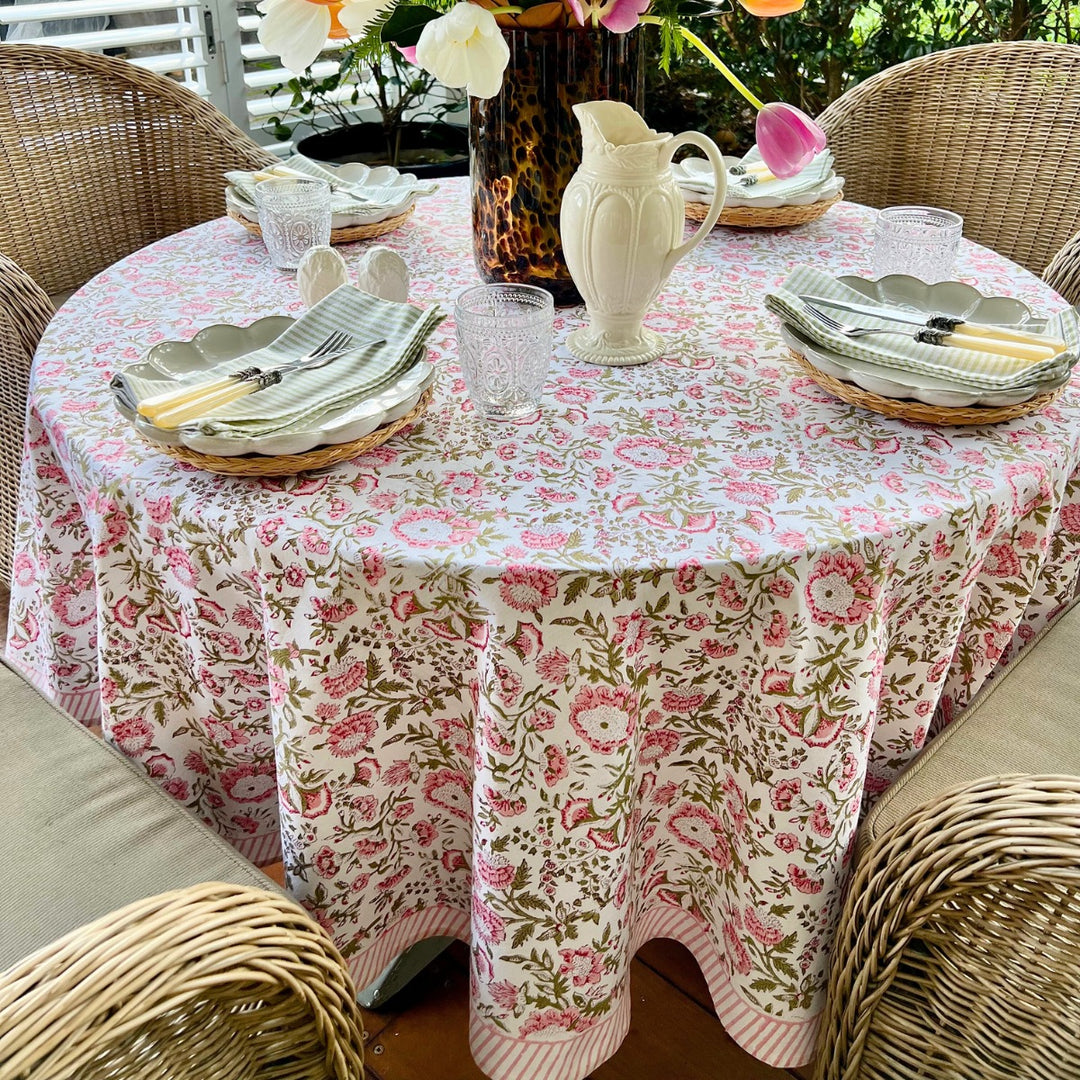 A round tablecloth with a floral pattern in shades of pink and green, displayed on a table set with dishes and a floral arrangement.