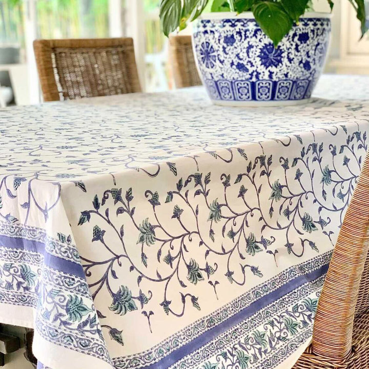 A square hand block print tablecloth with a blue green floral pattern and green vines on cotton fabric, laid out on a table with a cake and decorative pillow in the background.