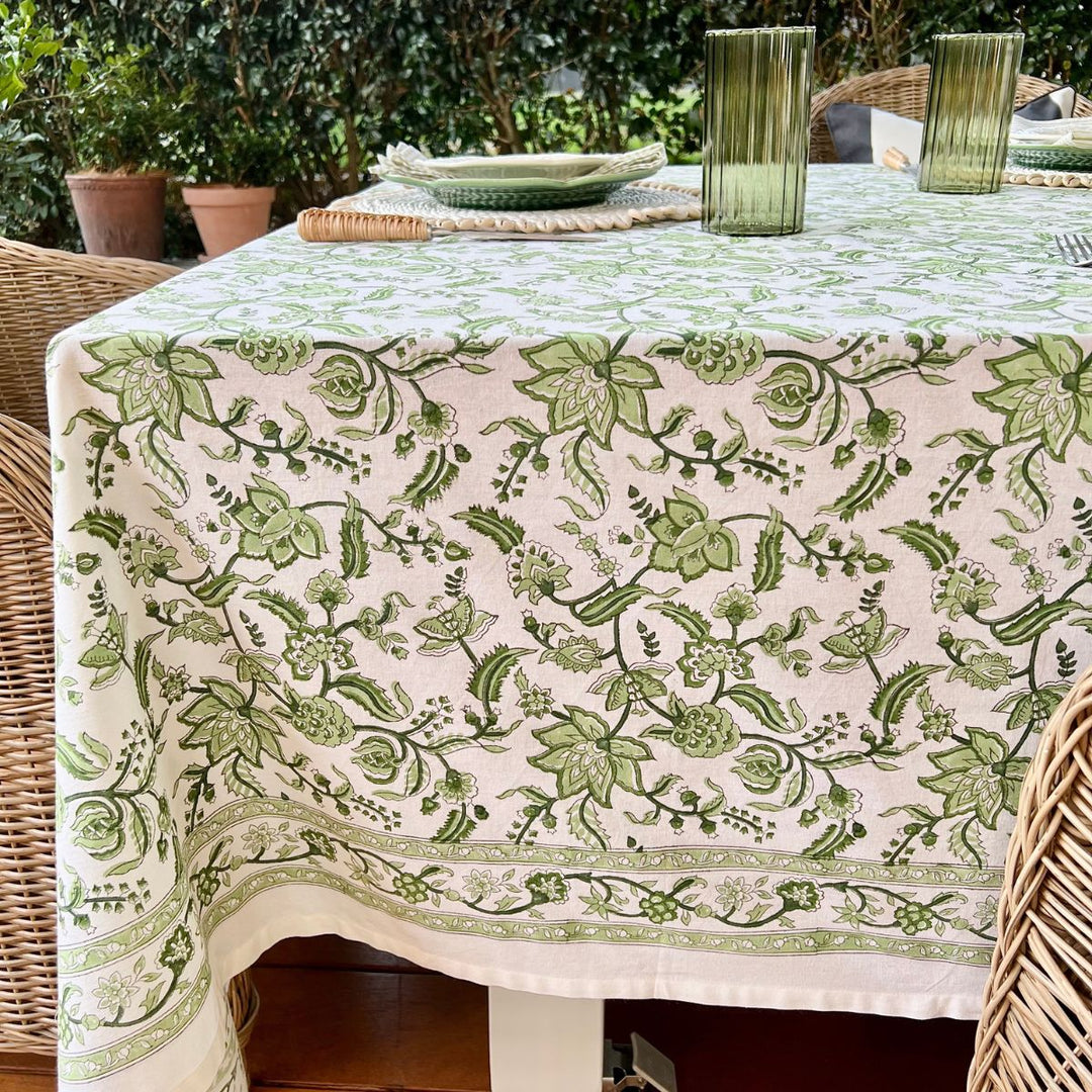 A green and white hand block printed chintz patterned square tablecloth displayed over a wicker table.