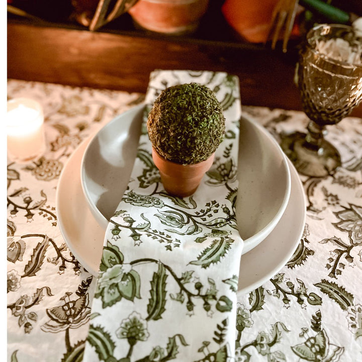 A green and white floral hand block printed patterned napkin is displayed on a white plate with a green plant-like decoration on top, set against a table with a floral tablecloth.