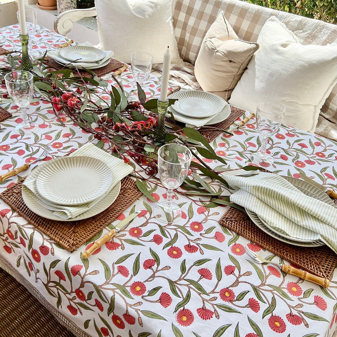A square tablecloth with a hand block printed  red flowering gum pattern, laid out on a dining table with plates, glasses, and napkins arranged on top.