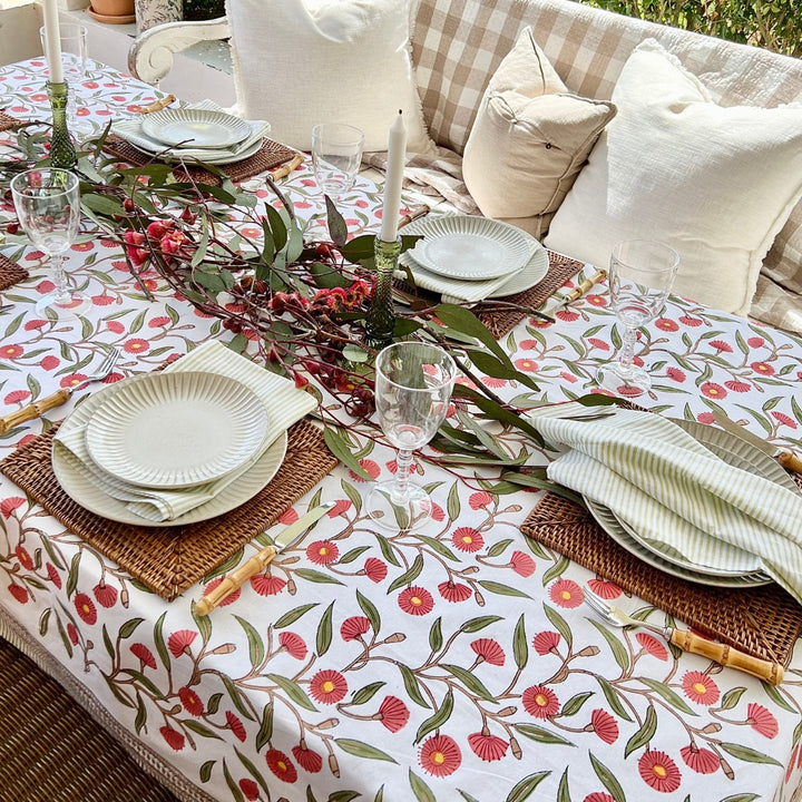 A square tablecloth with a hand block printed  red flowering gum pattern, laid out on a dining table with plates, glasses, and napkins arranged on top.
