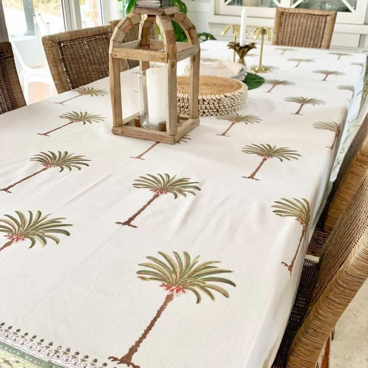 A square tablecloth with a green palm tree pattern, laid out on a table with a woven tablecloth border and a decorative lamp on top.