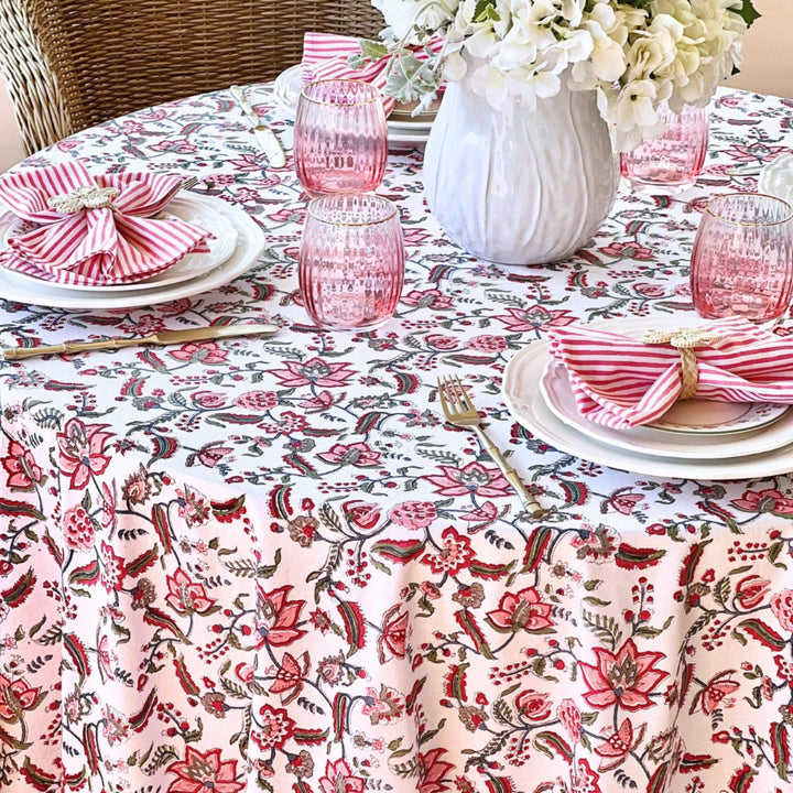 A round multicolored tablecloth with a floral pattern, featuring red and green leaves and pomegranate accents, displayed on a table with cutlery and a decorative vase.