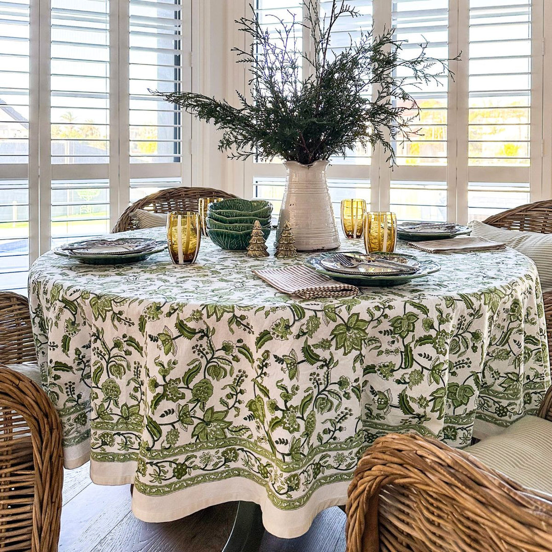 90 inch round tablecloth hand block printed tablecloth on a circular dining table, set with white green ceramic plates, amber glass tumblers at each place, and a loose arrangement of fresh greenery in a vase at the centre.
