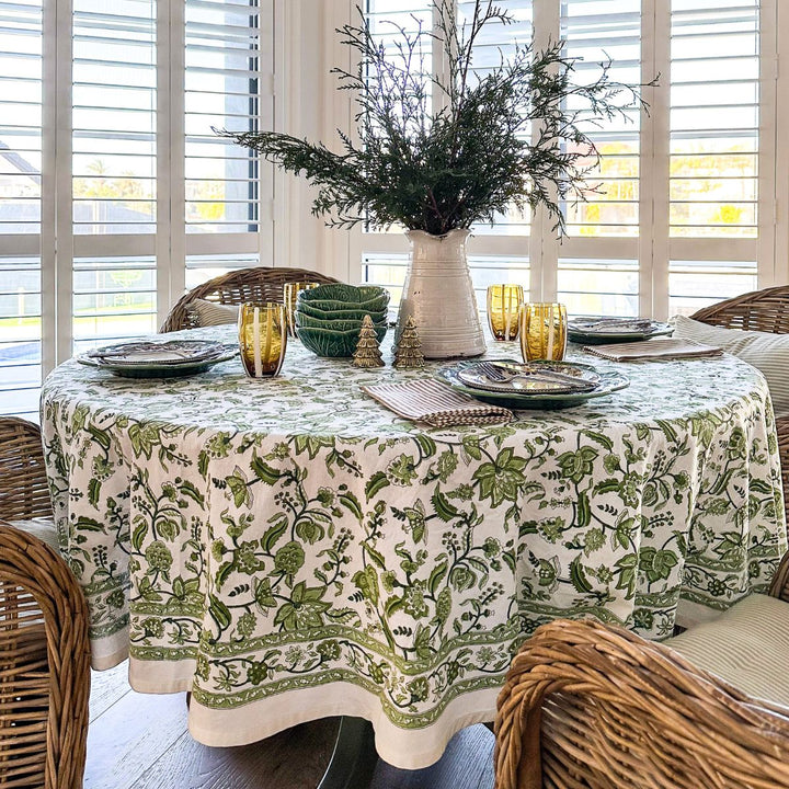 90 inch round tablecloth hand block printed tablecloth on a circular dining table, set with white green ceramic plates, amber glass tumblers at each place, and a loose arrangement of fresh greenery in a vase at the centre.