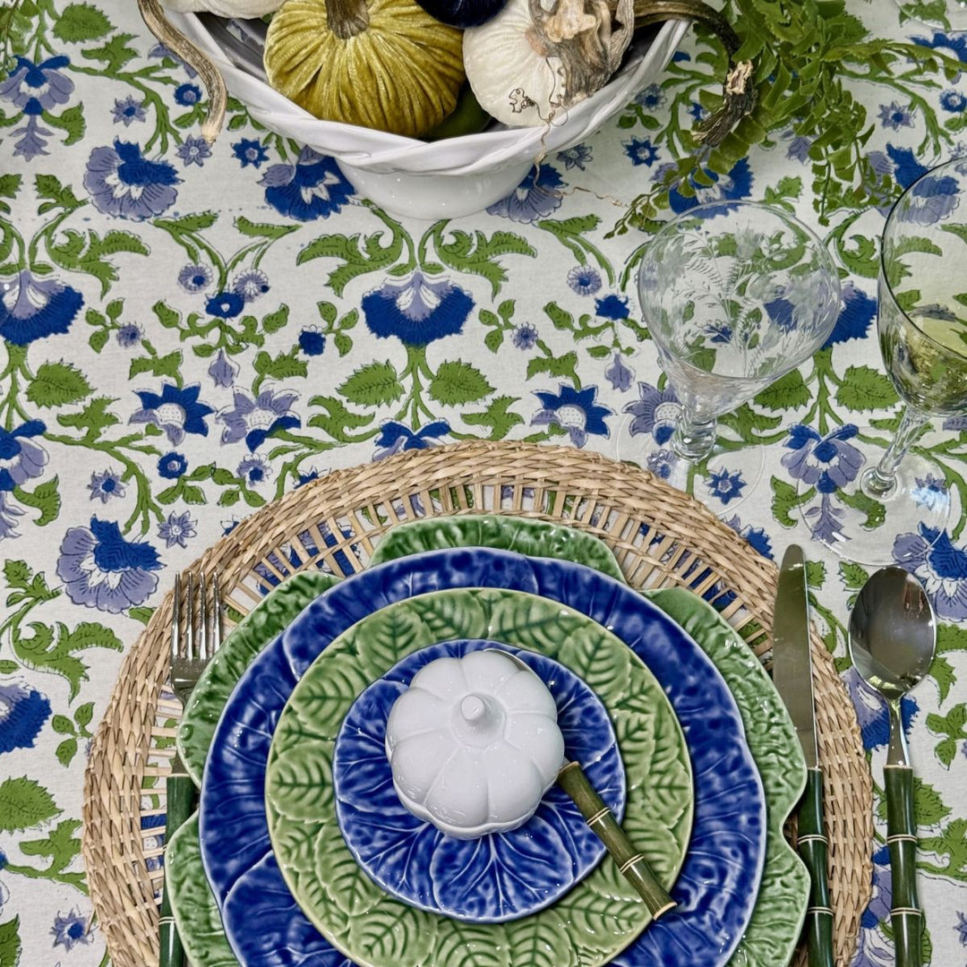 Close-up of a blue and green floral tablecloth with a woven placemat, layered dinner plates, and a white pumpkin for seasonal table styling.