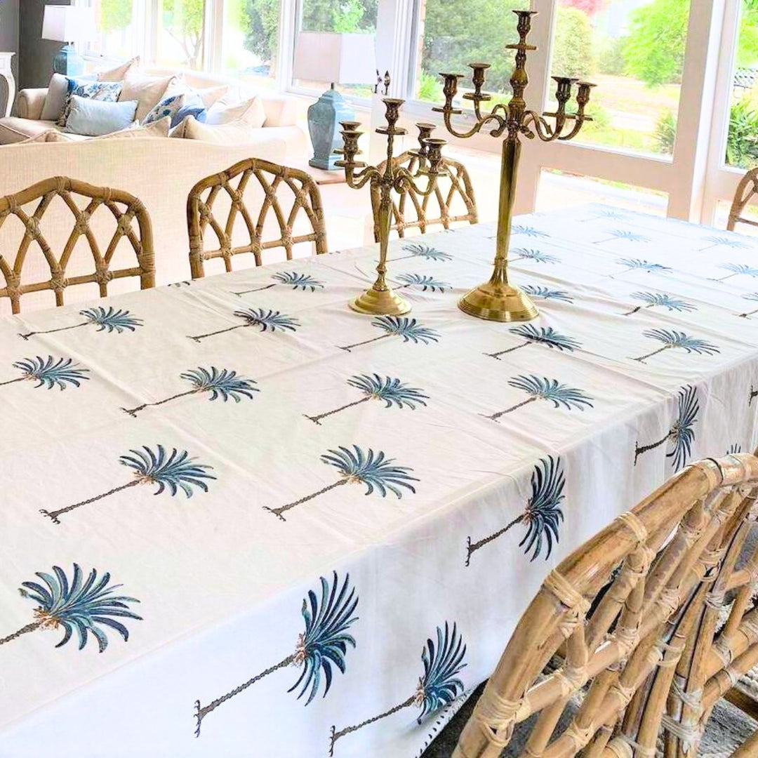 Corner of a square blue palm hand block printed tablecloth, showing the detailed hand block print on premium cotton, with  bamboo chairs and metal candlebaras with golden finish
