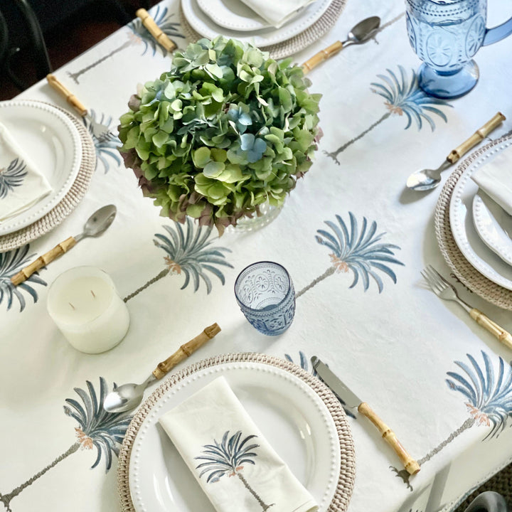 A casual weekend lunch on a rectangular blue palm hand block printed tablecloth with wicker chargers, white ceramic plates, bamboo cutlery, blue palm napkins knotted loosely beside each setting, and a low vase of green hydrangeas at the centre.