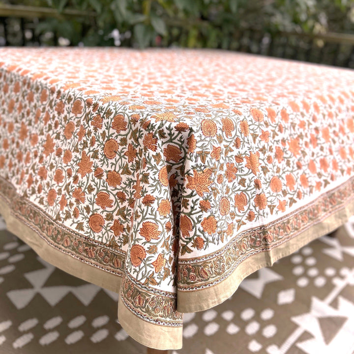 Corner of a rectangular warm rust and cream floral block printed tablecloth in 72 x 135 inches showing the layered border and tan trim, a fall table that seats 10 to 12