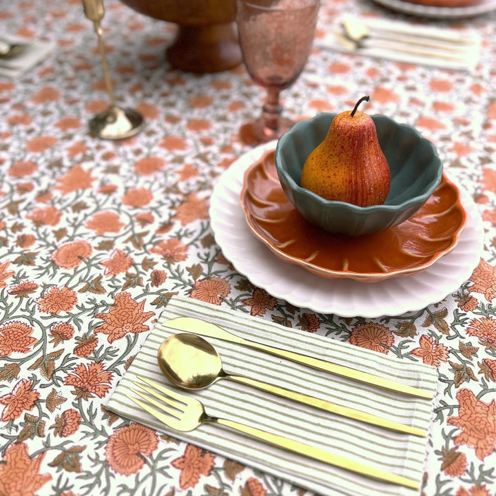 Close up of a harvest floral hand block printed tablecloth set for Thanksgiving with a scalloped rust plate, sage green bowl, fresh pear and gold cutlery