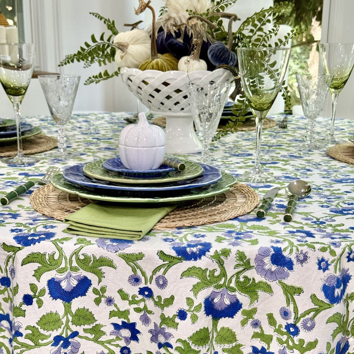 Blue and green floral round tablecloth draped over a dining table with layered blue-green plates, crystal glasses, and a white pumpkin centerpiece.