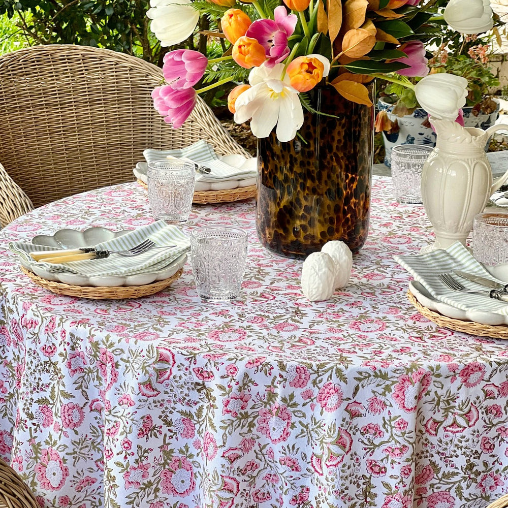 A floral round tablecloth with pink and green tones displayed on a table set for dining, with a vase of flowers and striped napkins complementing the tablecloth.