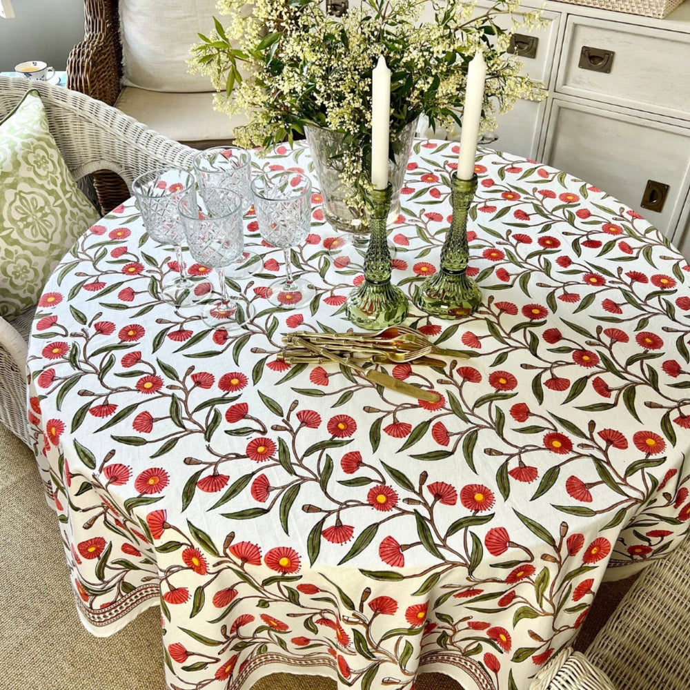 A round tablecloth with a red green flowering gum hand block printed design displayed on a table, paired with glassware and a white wicker chair in the setting.