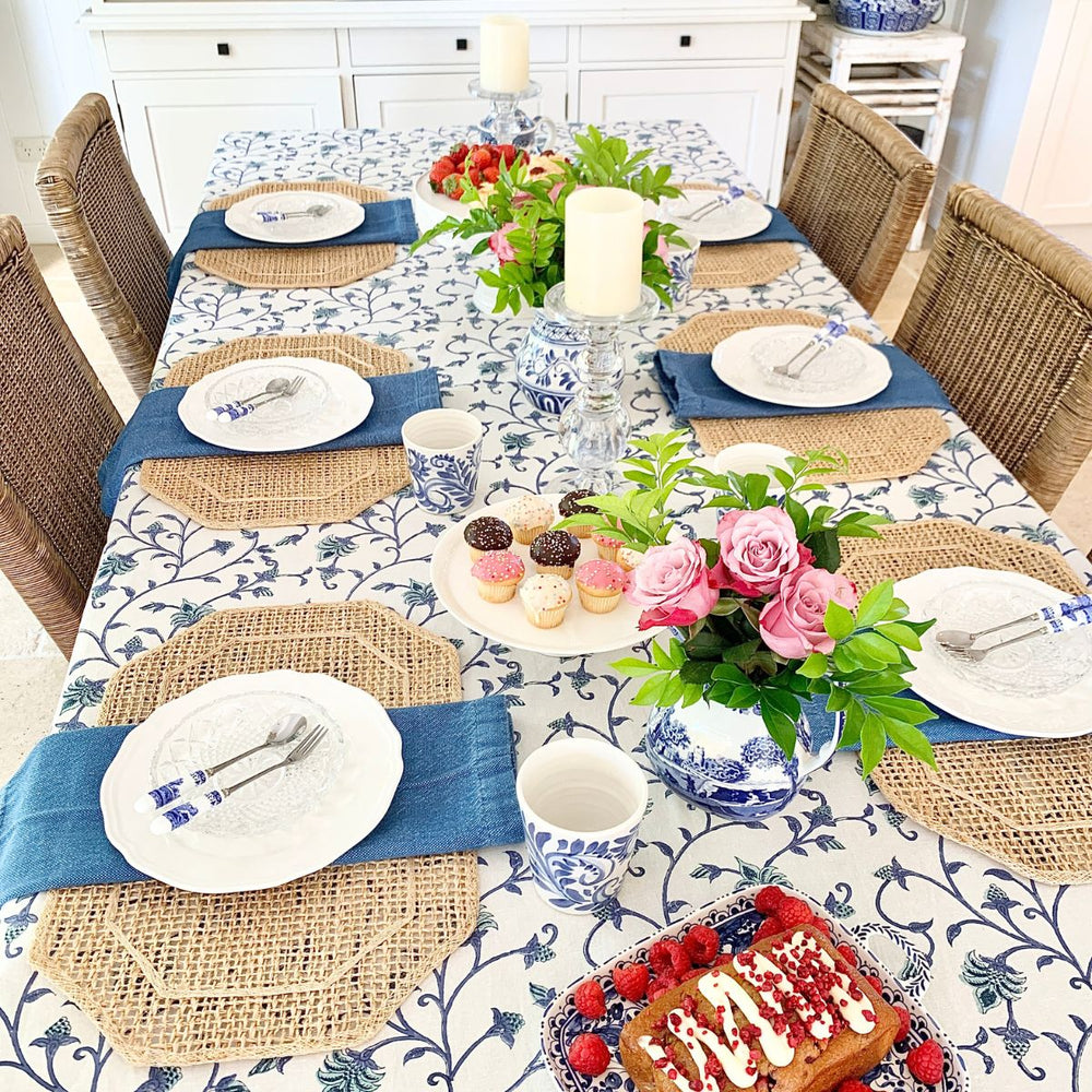 A square blue and green Ivy hand block printed cotton tablecloth on a four-seater dining table, set with white ceramic plates, glass tumblers, and a floral vase centrepiece.