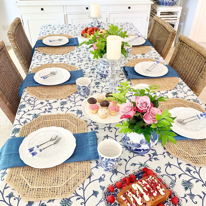 A square blue and green Ivy hand block printed cotton tablecloth on a four-seater dining table, set with white ceramic plates, glass tumblers, and a floral vase centrepiece.