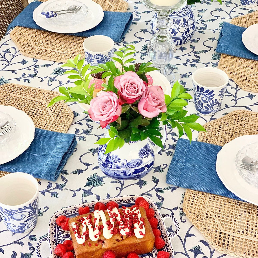A 70 inch square blue and green floral hand block printed tablecloth on a wooden dining table for four, styled with white dishes, folded napkins at each place, and a low floral vase arrangement as the centrepiece.