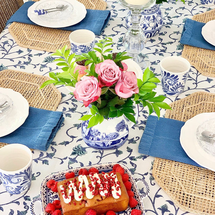 A 70 inch square blue and green floral hand block printed tablecloth on a wooden dining table for four, styled with white dishes, folded napkins at each place, and a low floral vase arrangement as the centrepiece.