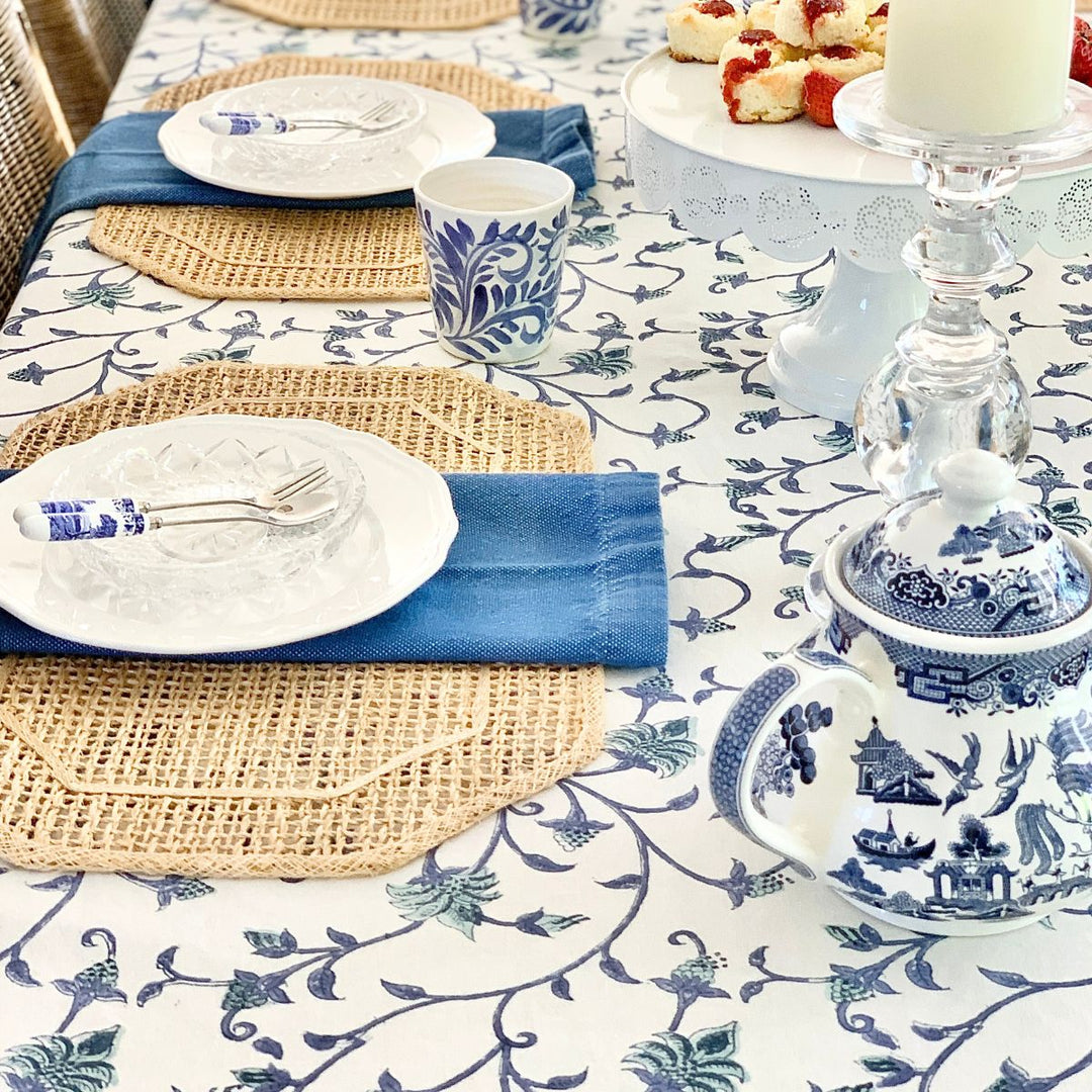 Close-up corner of a square blue and green floral hand block printed cotton tablecloth on a dining table, with a white plate and neatly placed cutlery in the foreground.
