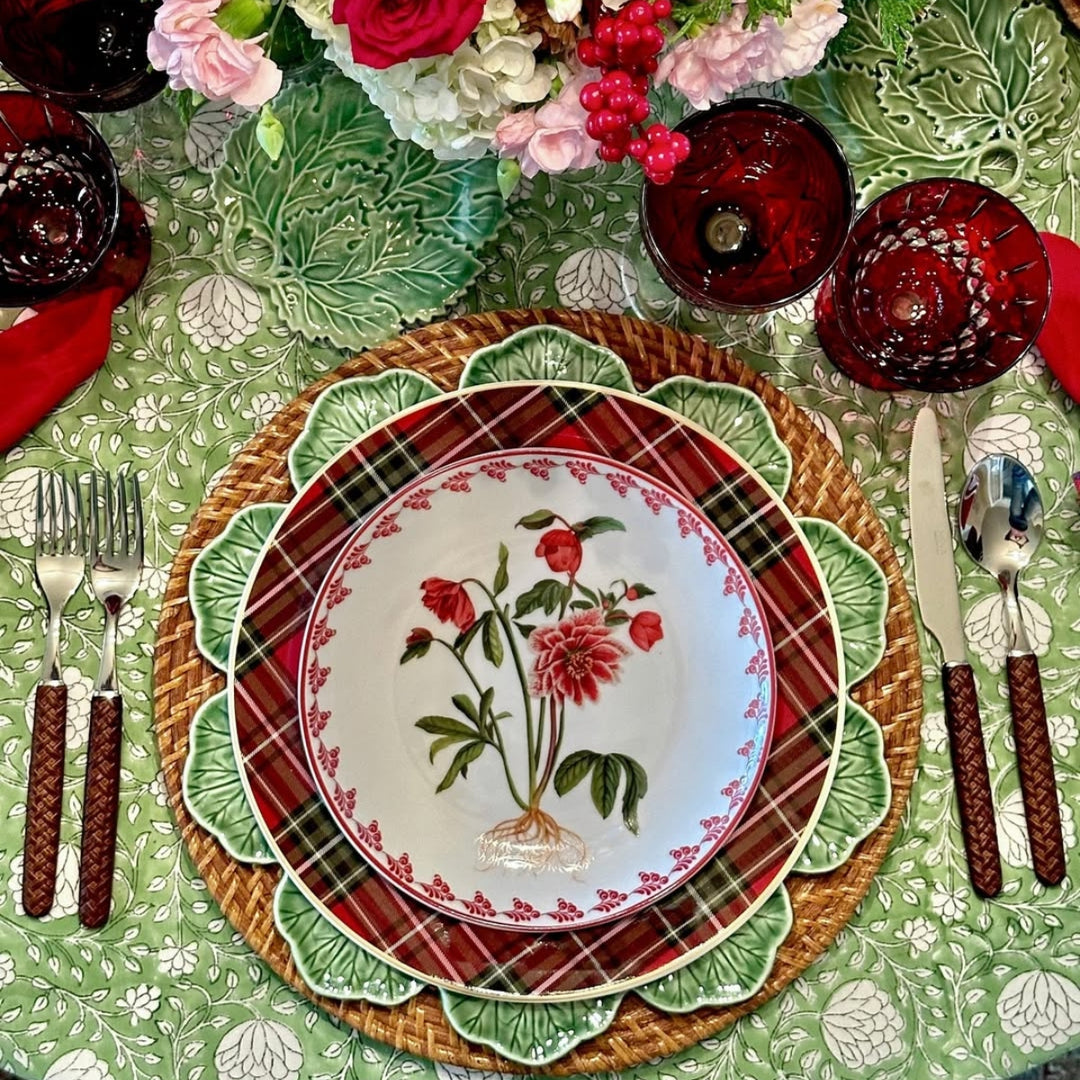 A round Eden tablecloth in green and white floral motif as the base for an intimate Christmas dinner, with a floral centrepiece, and red glassware. 