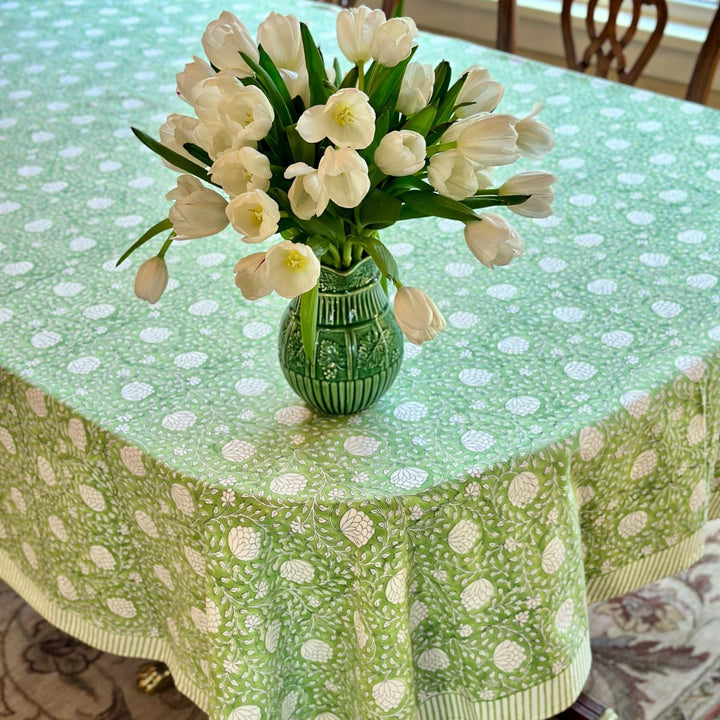 A green hand block printed rectangular tablecloth with a floral pattern displayed on a table with a vase of white flowers in the center.