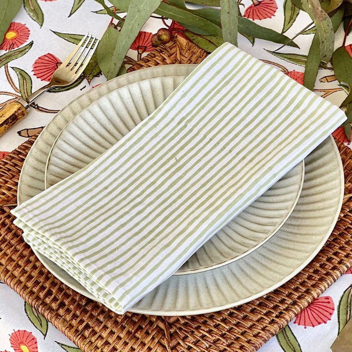 A set of green striped cotton napkins placed on a plate, with a second napkin partially visible in the background on a table with a floral tablecloth.