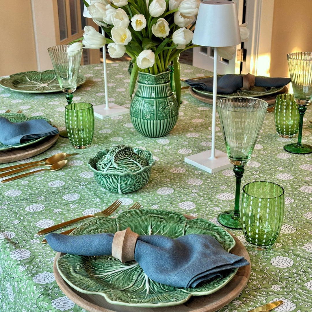 A square hand block printed Eden tablecloth in green and white on a large six-seater dining table, styled with cabbageware plates, a white floral centrepiece, and lamps. blue