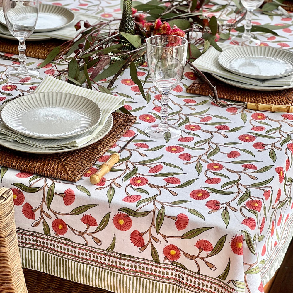 A square tablecloth with a red flowering gum pattern, laid out on a dining table with plates, glasses, and cutlery set on top.