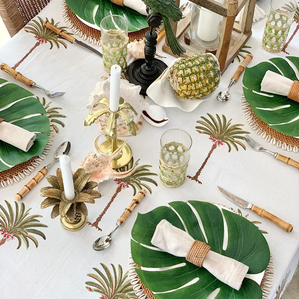 Top-down view of a hand block printed palm green tablecloth on a round dining table, featuring wicker chargers at each place, white dishes, bamboo cutlery, and napkins.