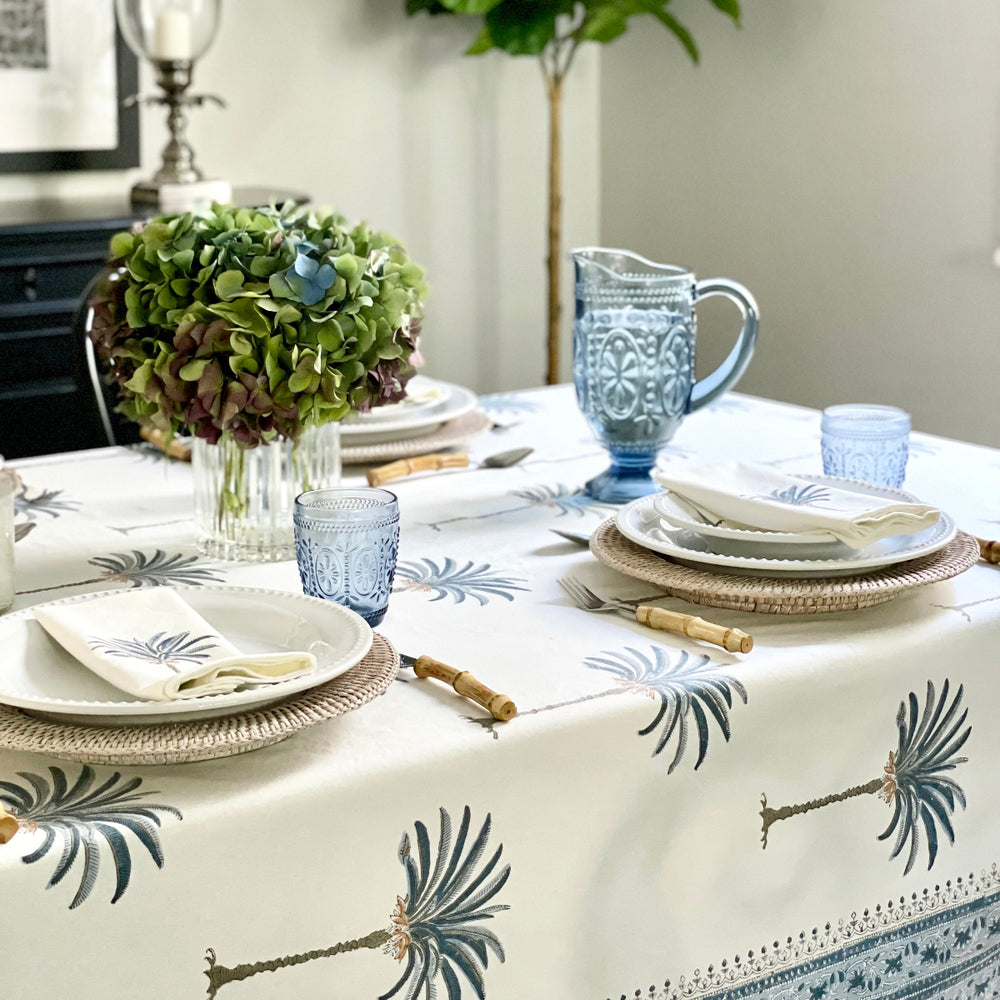 A bright casual table on a rectangular blue palm hand block printed tablecloth with an intricate border, with wicker chargers, white enamel dishes, bamboo cutlery, blue palm patterned napkins in rattan napkin rings, and fresh green stems in a simple vase.