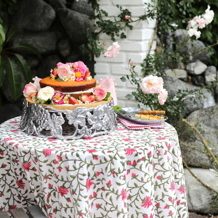 A beautiful outdoor garden table set on a 70 inch Pink green floral pattern round tablecloth with an intricate colourful border, styled with white plates, delicate glassware, and a stunning floral cake at the centre of the table.