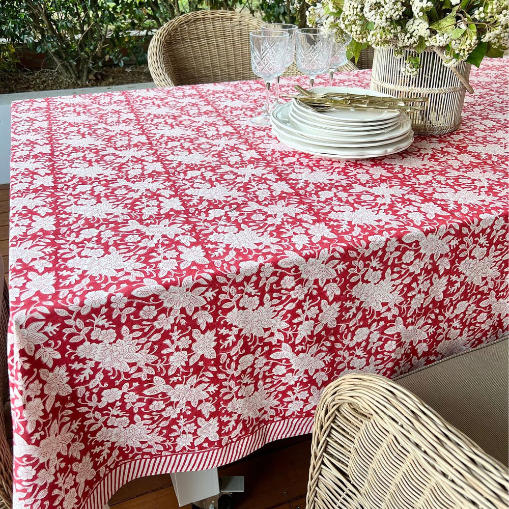 A red and white floral patterned square christmas tablecloth on a table with plates and cutlery set up for a meal.
