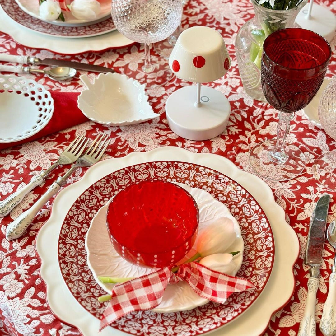 A circular 70 inch dining table covered with a round red and white hand block printed tablecloth, featuring white ceramic plates, a glass vase of fresh white flowers at the centre, and napkins folded simply beside each setting.