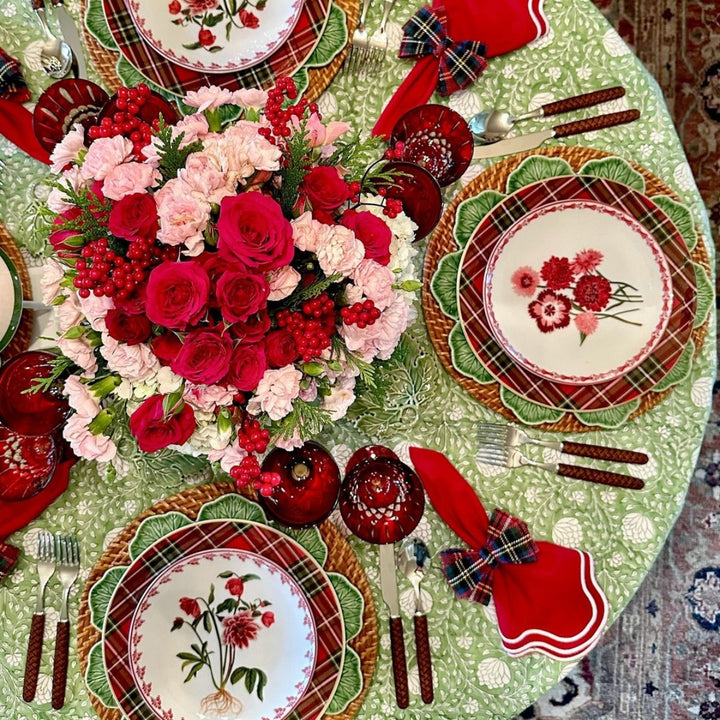 A round Eden tablecloth in green and white block print on a circular dining table dressed for Christmas, with a red and pink floral centrepiece, red glasses, and tapered candles. Red napkins folded with tartan check bows wth rattan chargers and cabbageware.