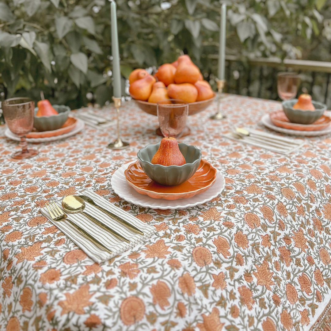 Rectangular orange and sage green floral hand block printed tablecloth set for a Thanksgiving table with pears, a harvest fruit bowl, taper candles and pink glassware