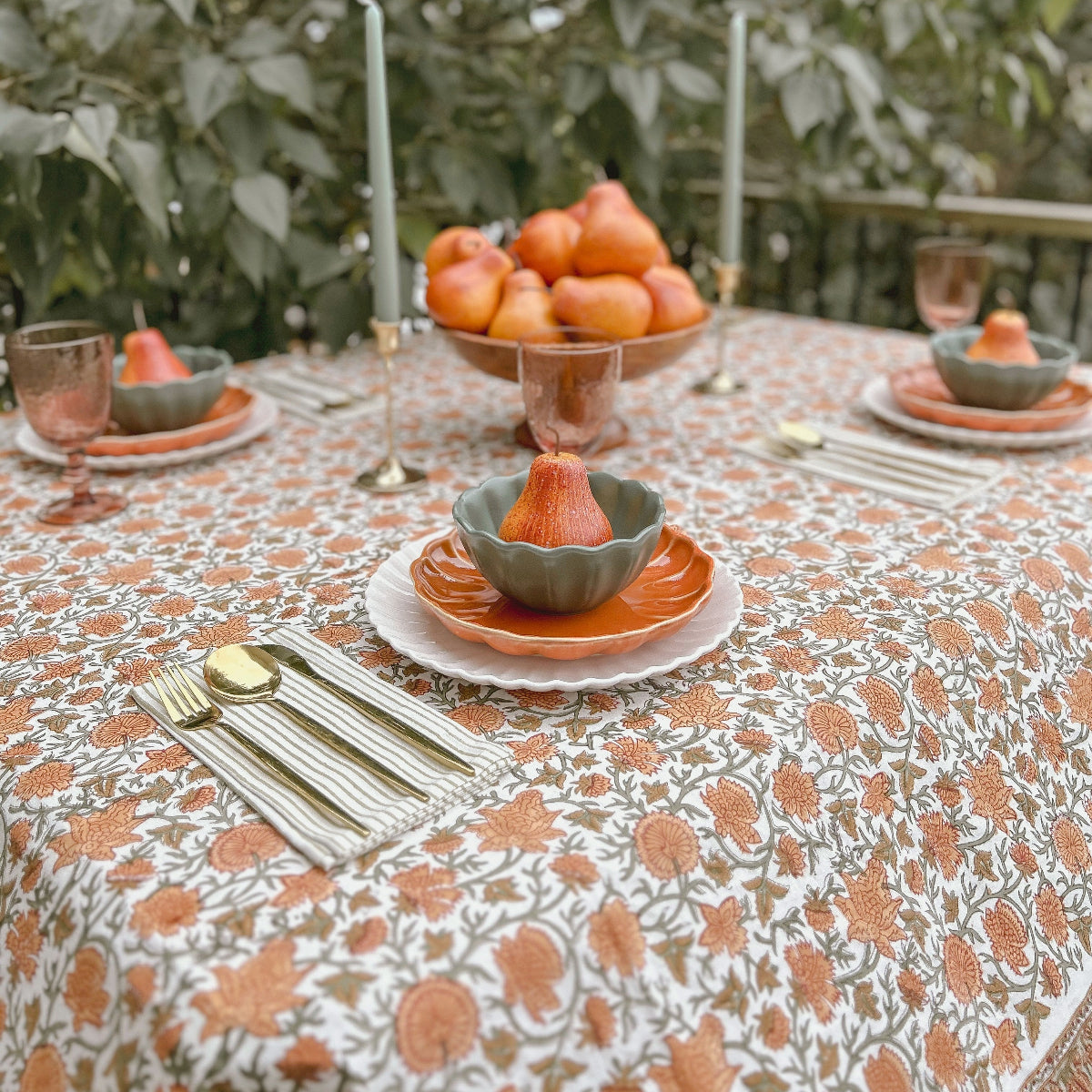 Rectangular orange and sage green floral hand block printed tablecloth set for a Thanksgiving table with pears, a harvest fruit bowl, taper candles and pink glassware