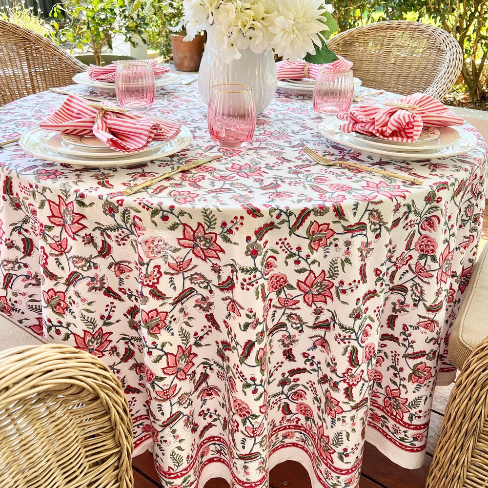 A multicolored round tablecloth with a pattern of hand block printed pomegranates and green leaves, displayed on a table set with plates and glasses.