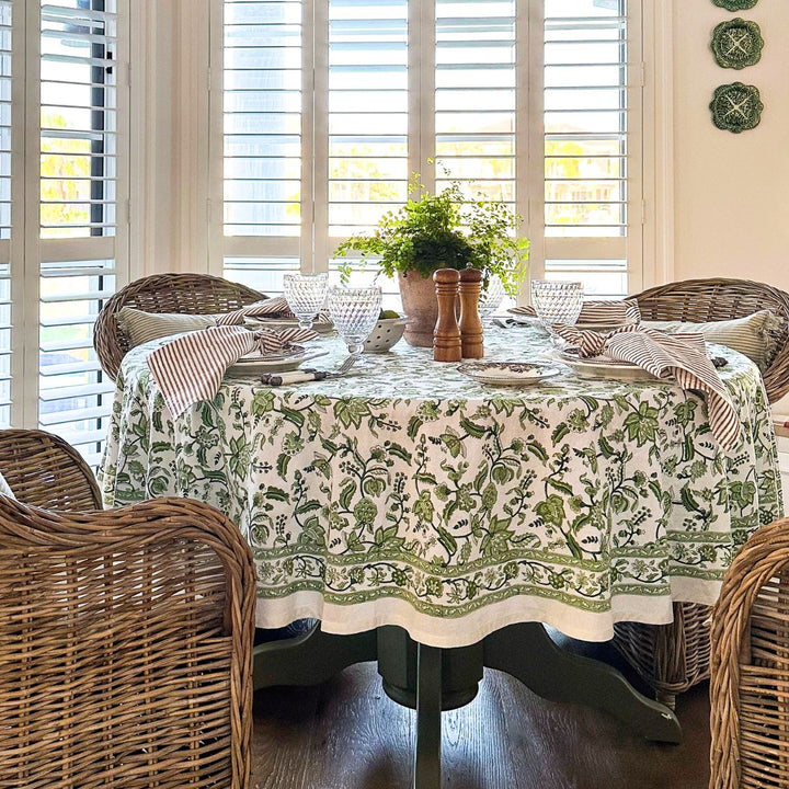 A casual cottagecore dining setup on a round Indian chintz floral hand block printed tablecloth, with a greenery centrepiece , vintage crockery, and clear glasses near large sunny windows.