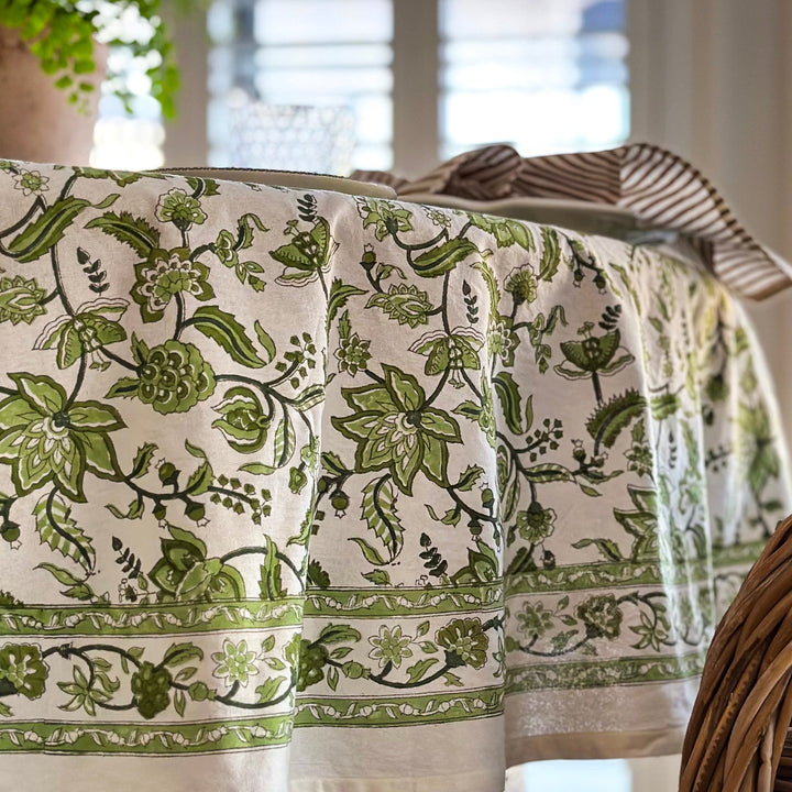 
A relaxed casual table on a round Indian chintz floral hand block printed tablecloth, with white dishes, amber glasses, and green foliage vase, fresh, simple, and effortlessly styled.