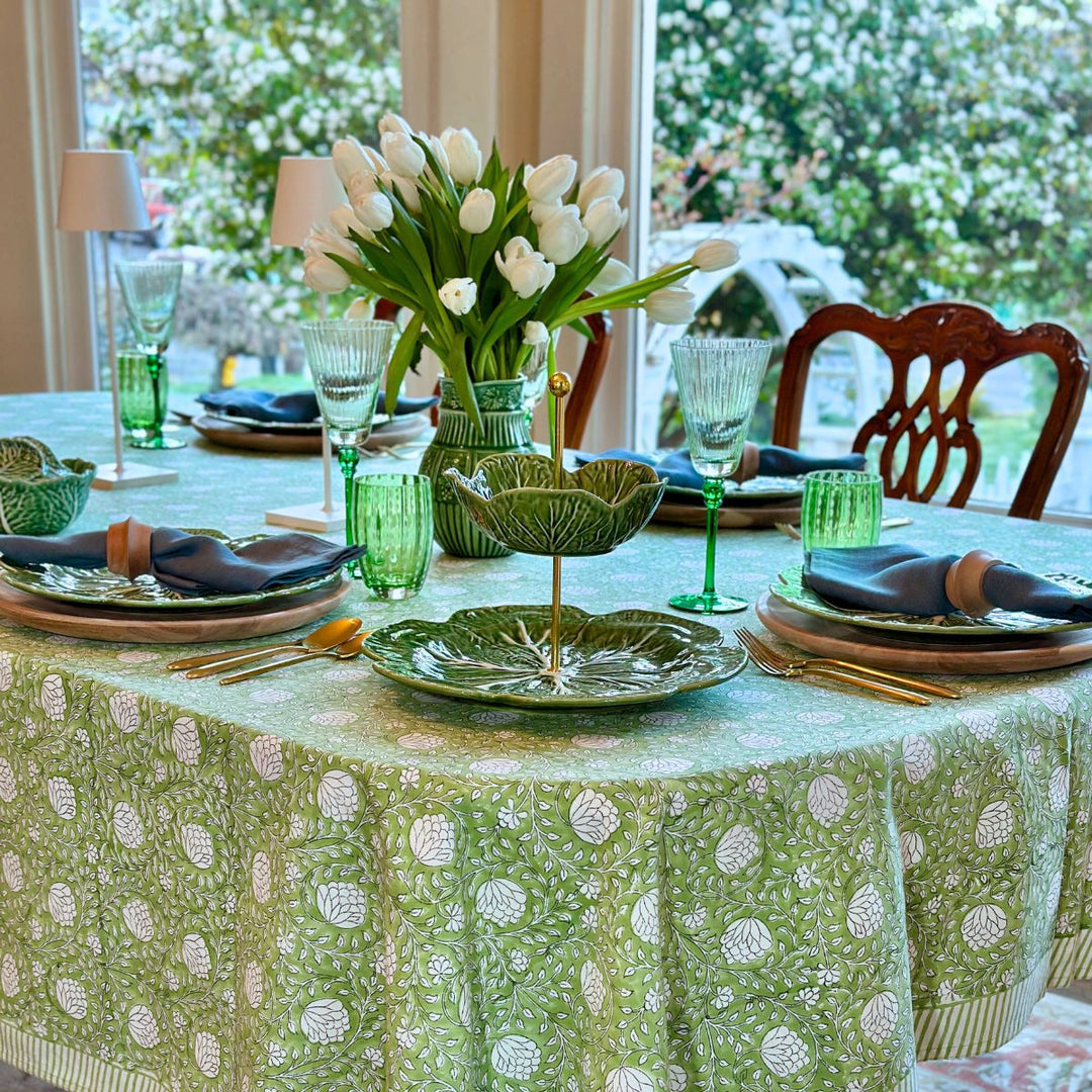 A rectangular hand block printed Eden tablecloth in green and white, laid on a long dining table with stacked green cabbageware plates and a centrepiece of fresh flowers with blue linen napkins folded beside each place setting.