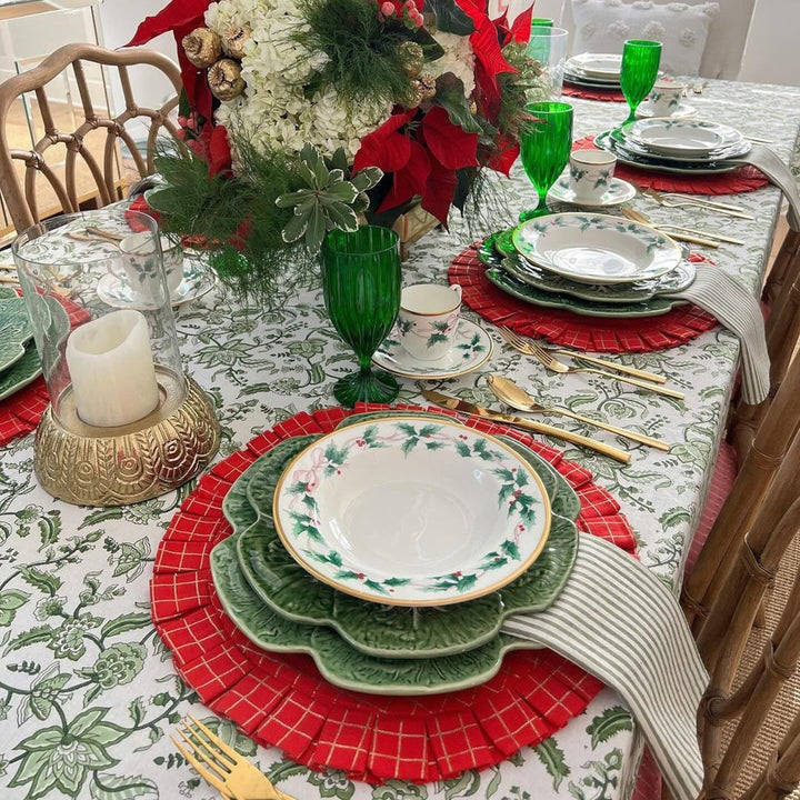 A rectangular hand block printed Indian chintz floral tablecloth on a long dining table dressed for Christmas, with green striped cotton napkins, festive dishes, lush green foliage in a vase, and delicate greenglass ornaments