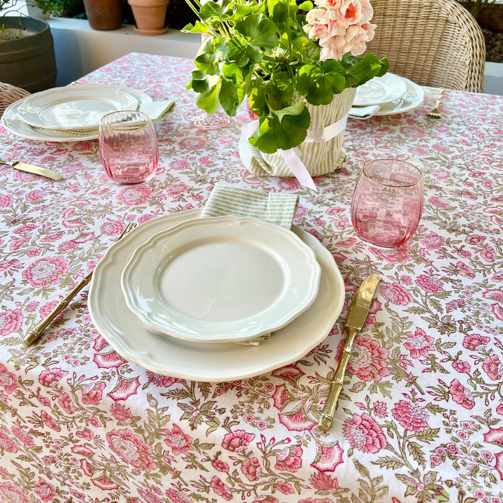 A rectangular block printed tablecloth featuring pink blooms and green foliage, laid on a six-seater table with brass cutlery and pink glass tumblers.