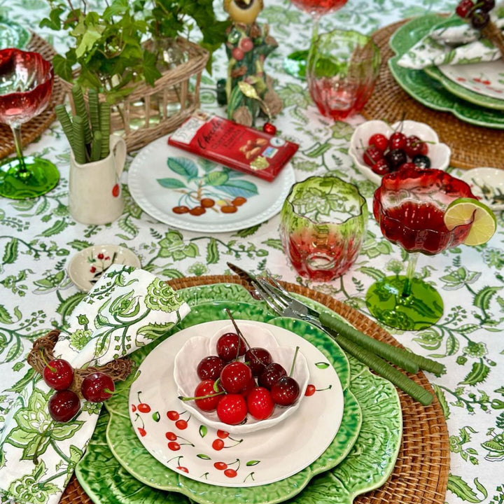 A cosy cottagecore table set with a round Indian chintz floral hand block printed tablecloth, featuring cabbageware dishes, cherries, greenery, and softly folded chintz pattern cotton napkins beside each place setting