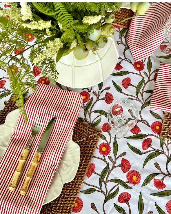 A set of red striped hand block printed table napkins with a floral pattern on the tablecloth, displayed alongside a glass of water and decorative greenery.