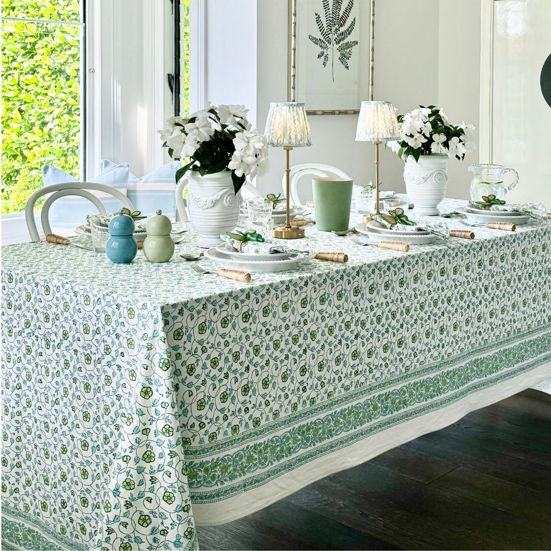 A square floral pattern tablecloth hand block printed in shades of green and blue, displayed on a table with dishes and decorative items.